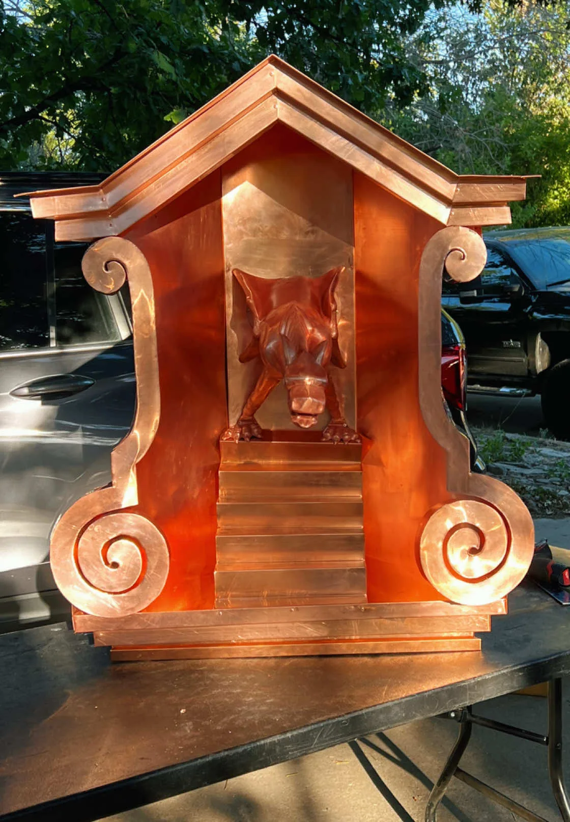 Fabricated copper pediment with scrolled brackets and a decorative face, standing on a bench outside the shop