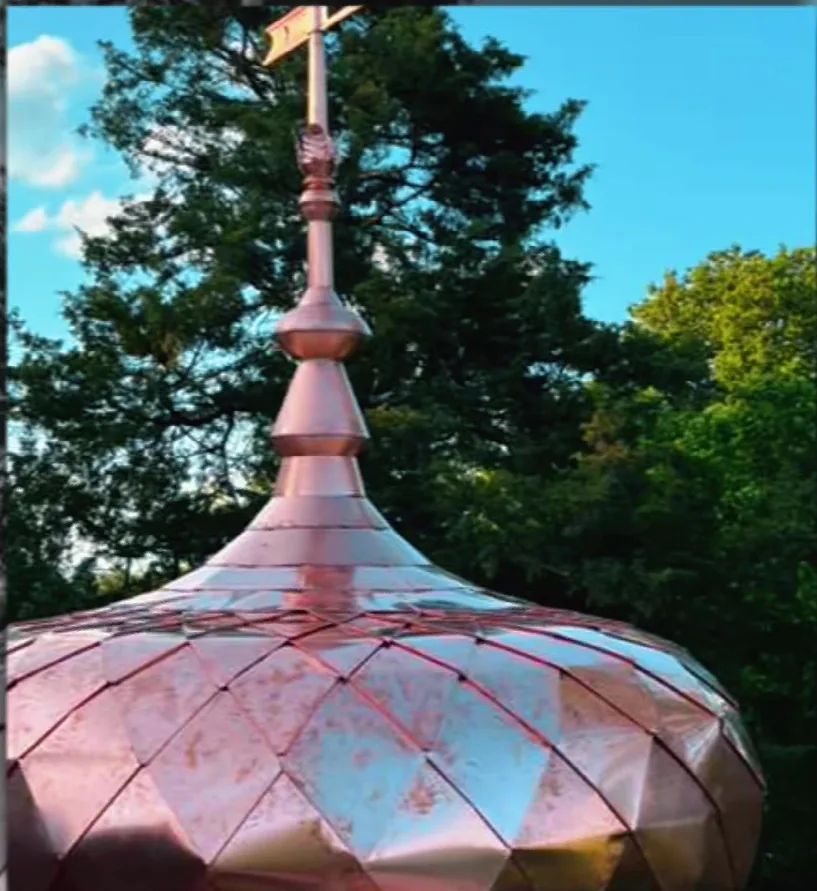Copper onion dome with diamond-pattern panels and a turned copper finial carrying a weather vane, new copper against trees and sky